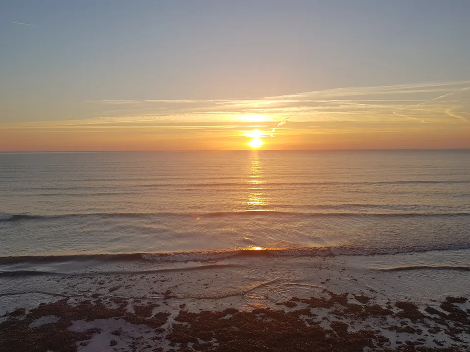 Orange sunlight stretches across the sea beneath a band of cloud as small waves reach the shore.