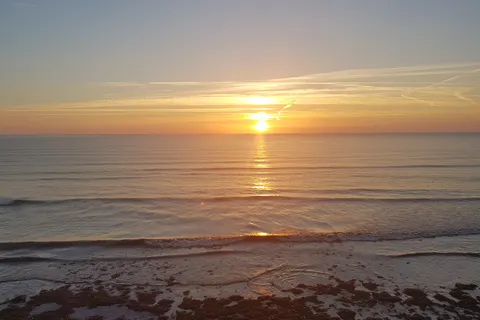 Orange sunlight stretches across the sea beneath a band of cloud as small waves reach the shore.