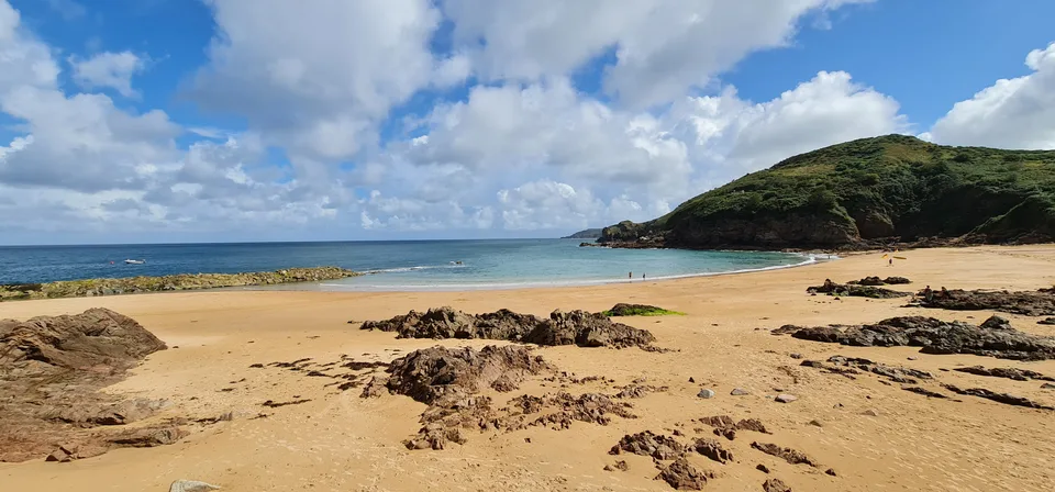 A wide view takes in golden sand, exposed rocks, blue water and a green headland at Grève de Lecq.