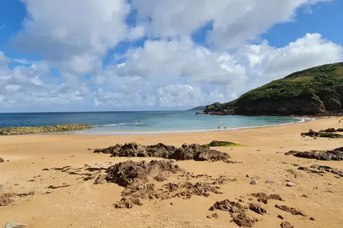 A wide view takes in golden sand, exposed rocks, blue water and a green headland at Grève de Lecq.