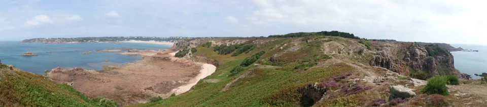 A wide panorama follows a rough coastal path across a rocky headland above blue sea beneath scattered clouds.