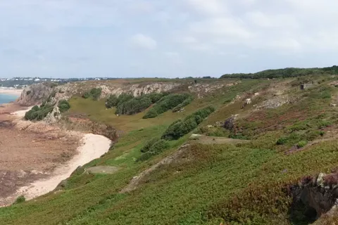 A wide panorama follows a rough coastal path across a rocky headland above blue sea beneath scattered clouds.