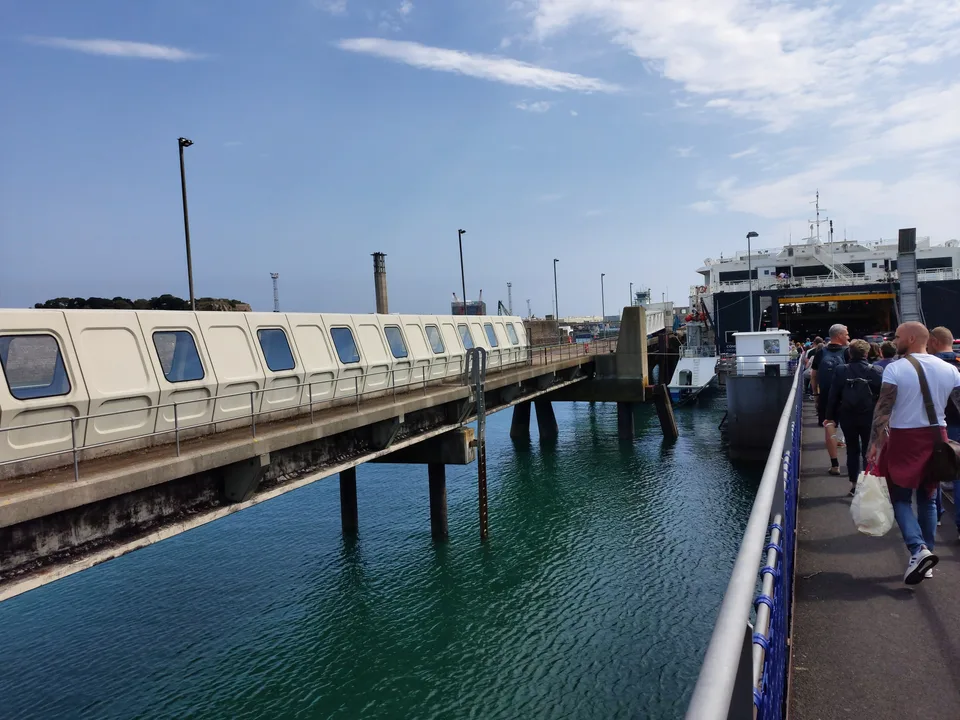 Passengers walk along a quayside towards a ferry beside blue harbour water and a covered passenger gangway at St Helier.