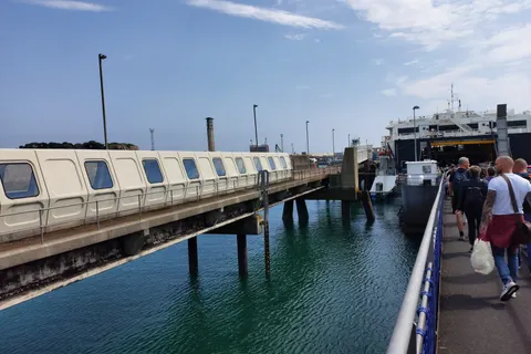 Passengers walk along a quayside towards a ferry beside blue harbour water and a covered passenger gangway at St Helier.