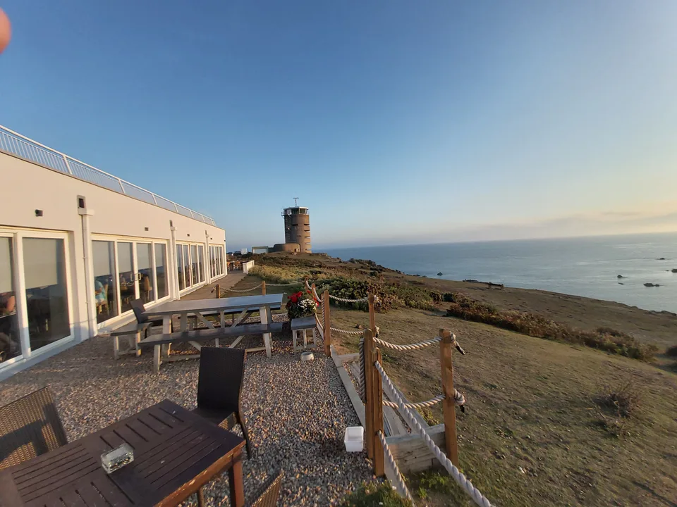 Wooden picnic tables and a rope fence stand beside a pale building above the rocky sea at La Corbière.