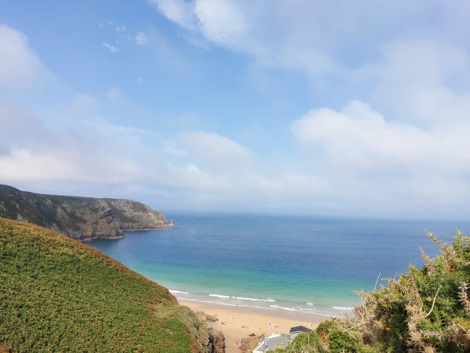 A green coastal hillside overlooks Plémont's sandy beach, turquoise shallows and a rocky headland beneath clouds.