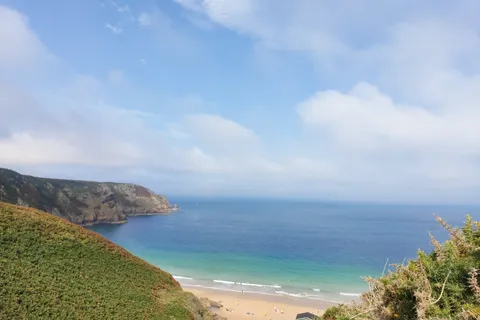 A green coastal hillside overlooks Plémont's sandy beach, turquoise shallows and a rocky headland beneath clouds.
