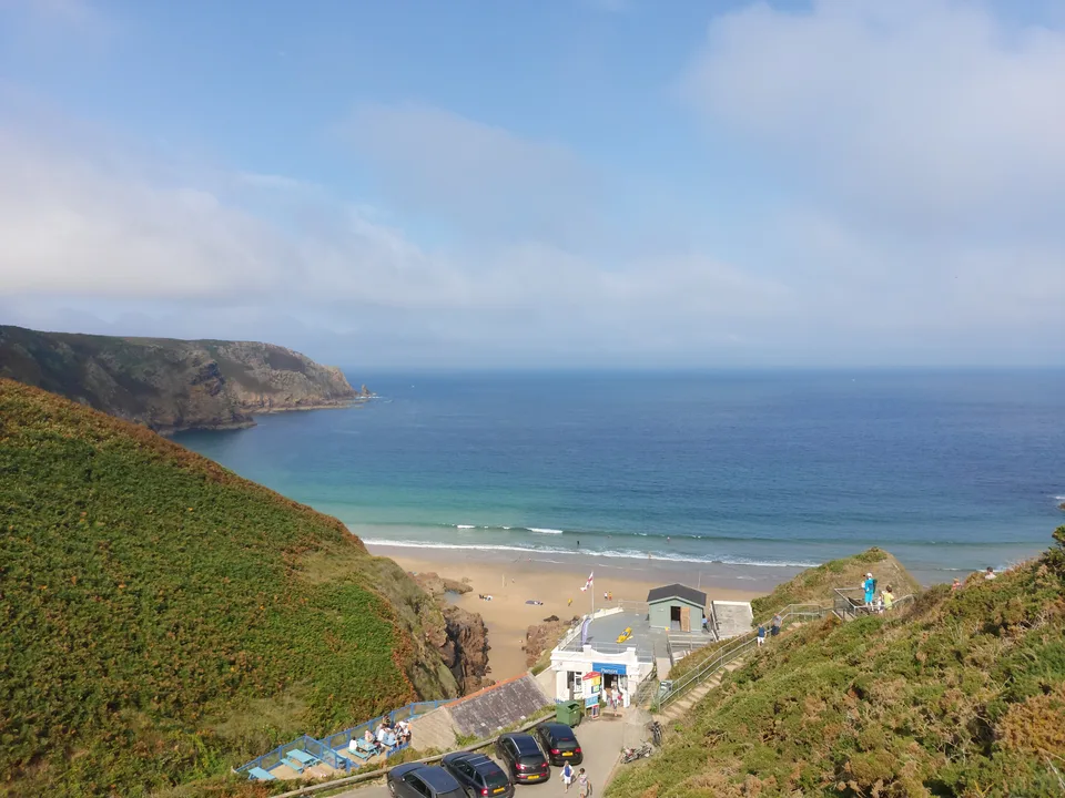 Small beach buildings sit between grassy cliffs and the sandy shore at Plémont, with blue sea beyond.