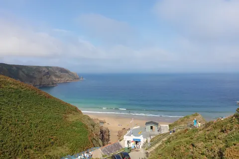 Small beach buildings sit between grassy cliffs and the sandy shore at Plémont, with blue sea beyond.