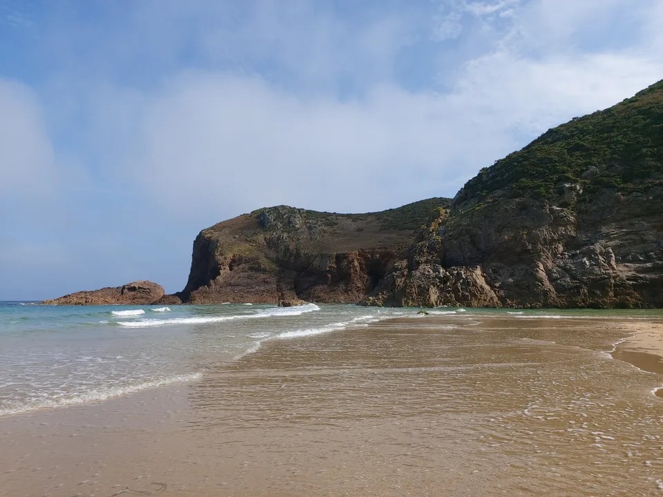 A rocky headland rises beside shallow waves and wet sand at Plémont under a partly cloudy sky.