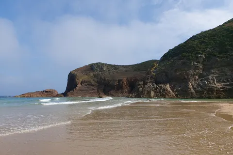 A rocky headland rises beside shallow waves and wet sand at Plémont under a partly cloudy sky.