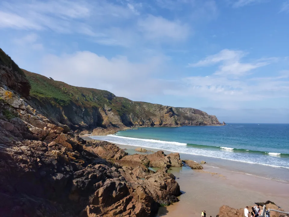 Dark foreground rocks frame Plémont's sandy cove, breaking waves and a distant green headland.