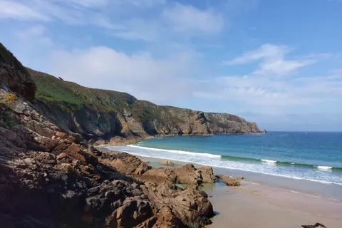 Dark foreground rocks frame Plémont's sandy cove, breaking waves and a distant green headland.