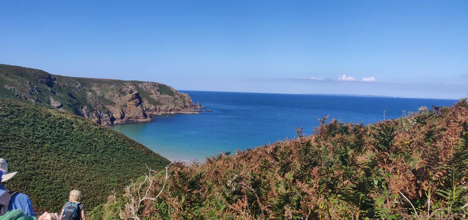 A wide view between grassy slopes reveals Plémont's sandy cove, rugged cliffs and blue sea under a clear sky.