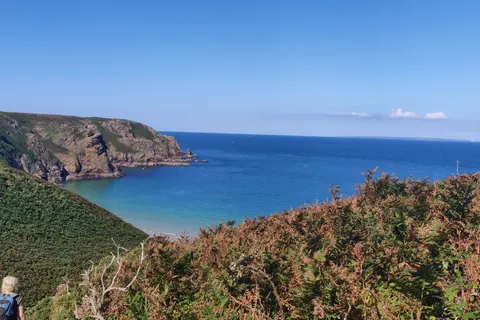 A wide view between grassy slopes reveals Plémont's sandy cove, rugged cliffs and blue sea under a clear sky.