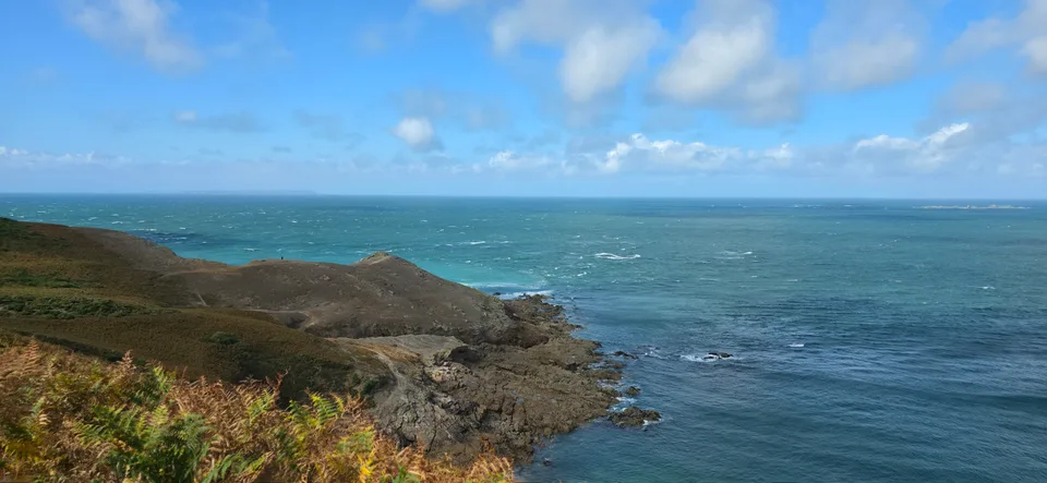 Low coastal vegetation and a rocky headland overlook a choppy blue sea beneath scattered clouds at Plémont.