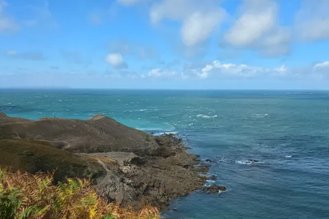 Low coastal vegetation and a rocky headland overlook a choppy blue sea beneath scattered clouds at Plémont.