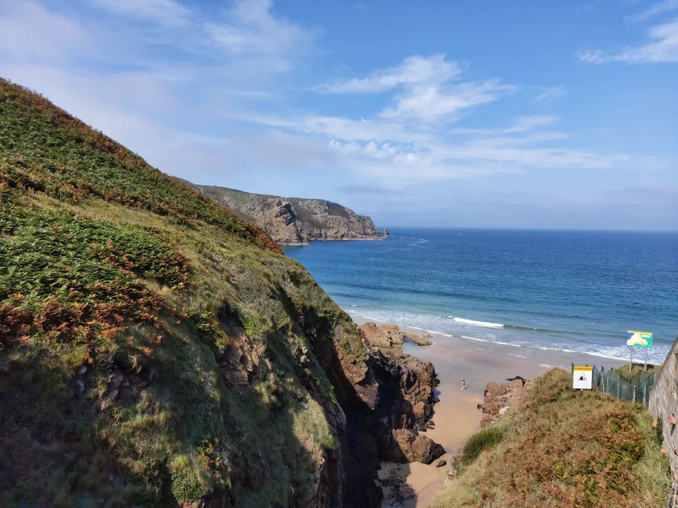 Green coastal cliffs frame Plémont's beach access and pale sand beside blue water.