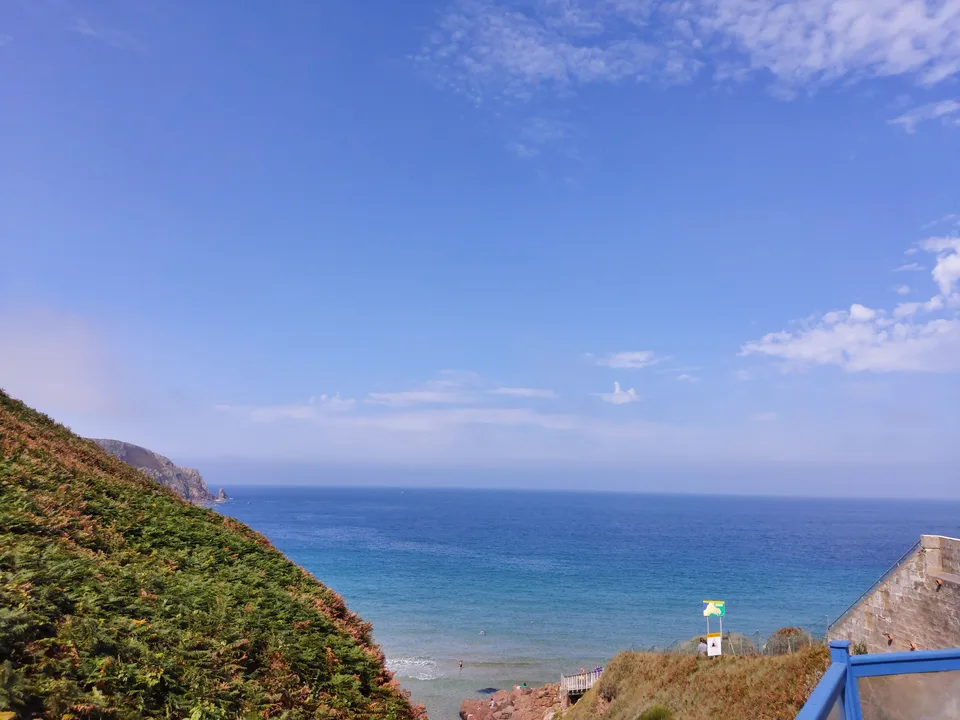 A green slope drops towards Plémont's shoreline, with blue sea and scattered clouds beyond a beach building.