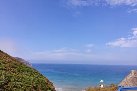 A green slope drops towards Plémont's shoreline, with blue sea and scattered clouds beyond a beach building.