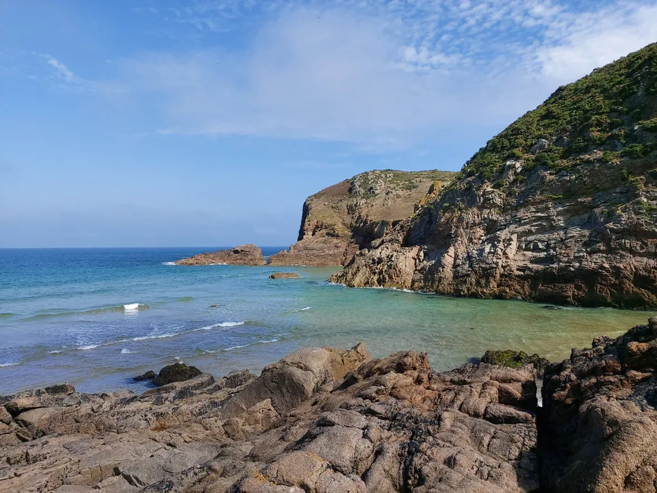 Dark layered rocks fill the foreground beneath a steep coastal cliff beside blue-green water at Plémont.
