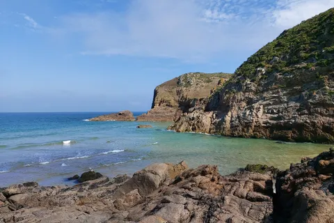 Dark layered rocks fill the foreground beneath a steep coastal cliff beside blue-green water at Plémont.