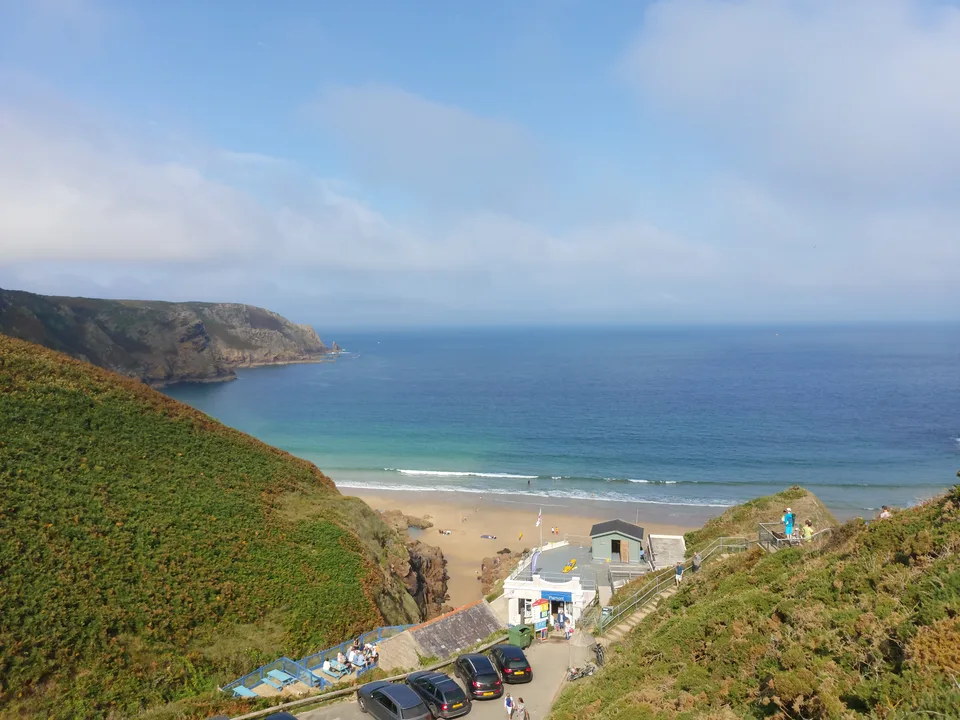 Grassy hillsides frame Plémont's sandy cove and small beach buildings beneath a partly cloudy sky.