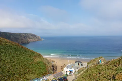 Grassy hillsides frame Plémont's sandy cove and small beach buildings beneath a partly cloudy sky.