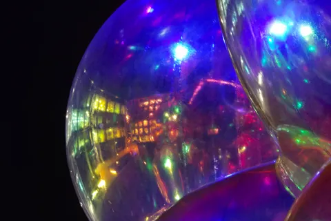 Purple, blue and white lights reflect across the curved surface of a transparent sphere beside a lit building.