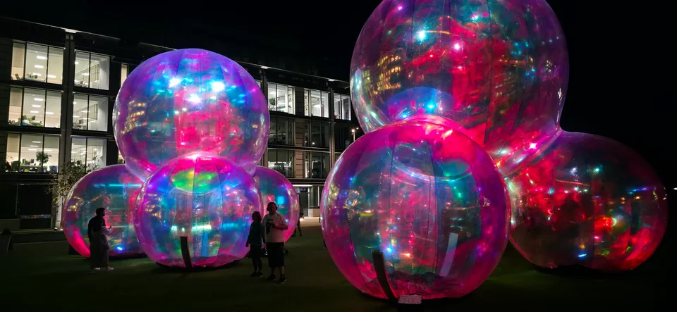 Large reflective spheres glow pink, blue and green beside illuminated windows against a dark night sky.