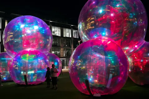 Large reflective spheres glow pink, blue and green beside illuminated windows against a dark night sky.