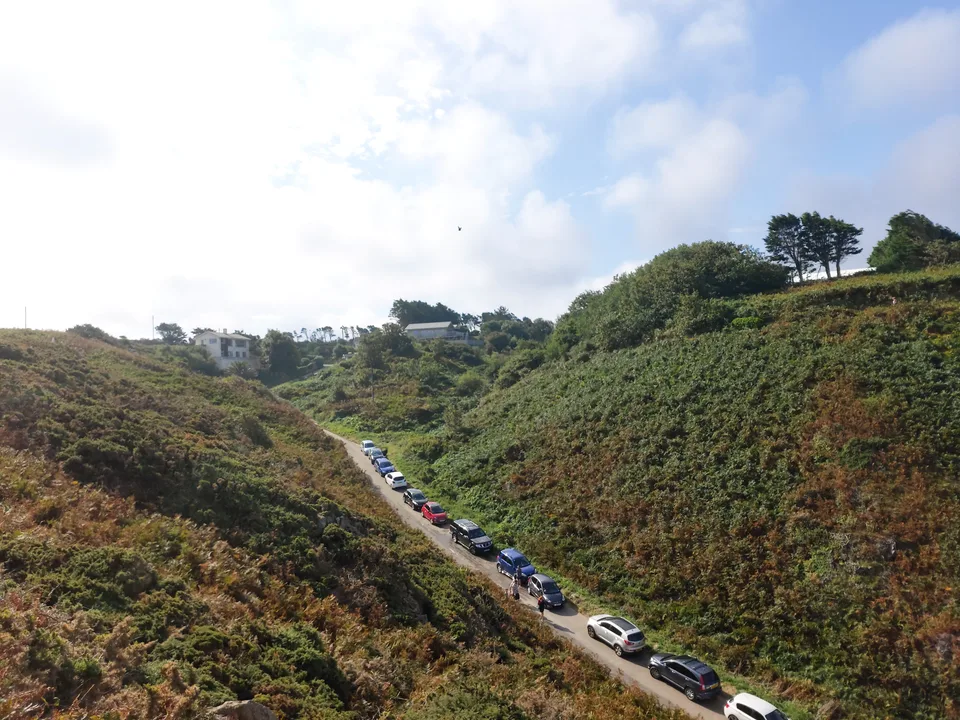 A narrow road lined with cars runs between green coastal hills above the beach at Plémont.