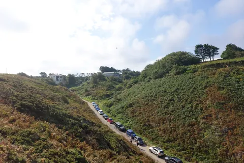A narrow road lined with cars runs between green coastal hills above the beach at Plémont.