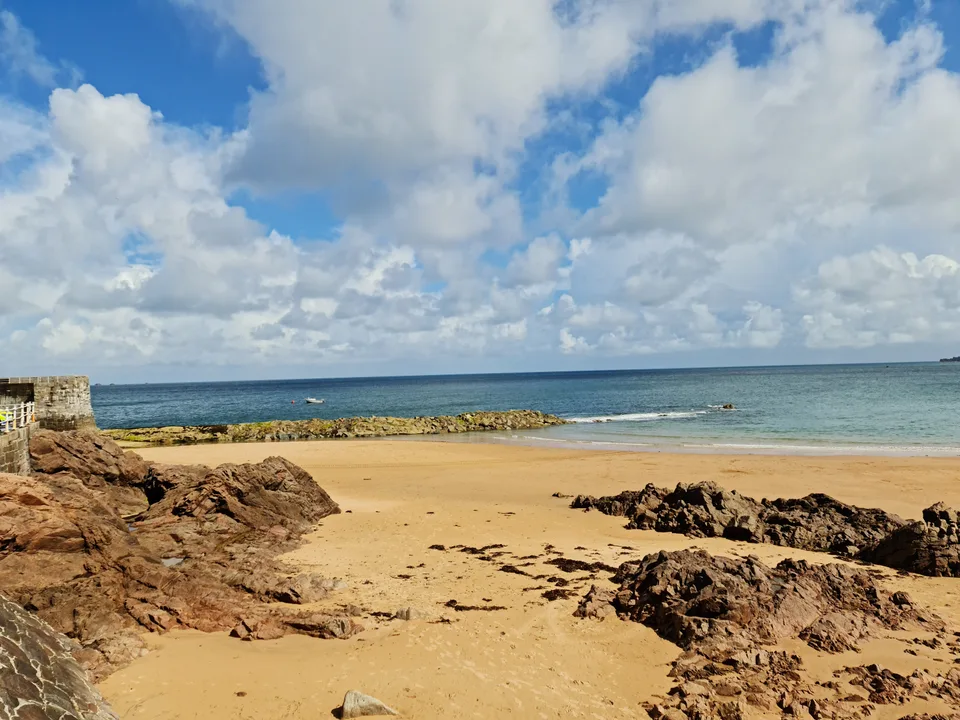 Low rocks emerge from golden sand beside calm sea and a stone harbour wall at Grève de Lecq under bright clouds.