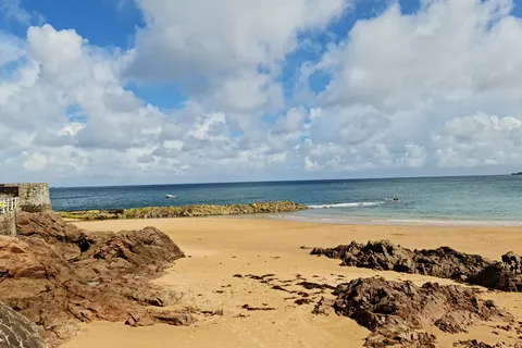 Low rocks emerge from golden sand beside calm sea and a stone harbour wall at Grève de Lecq under bright clouds.