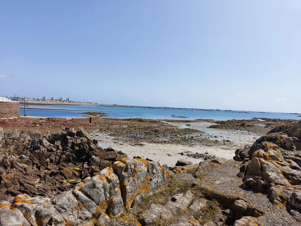 Dark rocks, seaweed and shallow tidal pools line the St Helier shoreline beneath a clear blue sky.