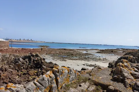 Dark rocks, seaweed and shallow tidal pools line the St Helier shoreline beneath a clear blue sky.