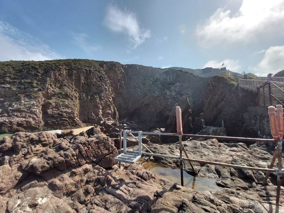 A metal railing crosses uneven shoreline rocks beneath a cliff at Plémont under a lightly clouded sky.