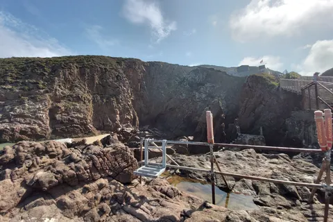 A metal railing crosses uneven shoreline rocks beneath a cliff at Plémont under a lightly clouded sky.