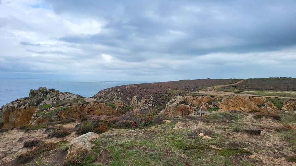 A rough path winds among low rocks and coastal grass above the sea at Grosnez beneath a grey-blue sky.