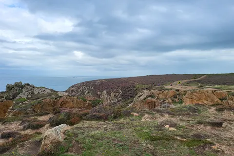 A rough path winds among low rocks and coastal grass above the sea at Grosnez beneath a grey-blue sky.