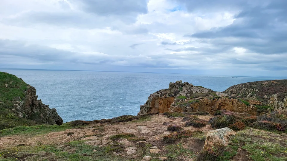 Uneven rocks and sparse coastal grass overlook a blue-grey sea from the headland at Grosnez beneath clouds.