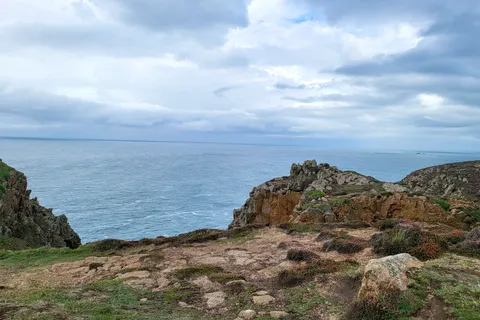 Uneven rocks and sparse coastal grass overlook a blue-grey sea from the headland at Grosnez beneath clouds.