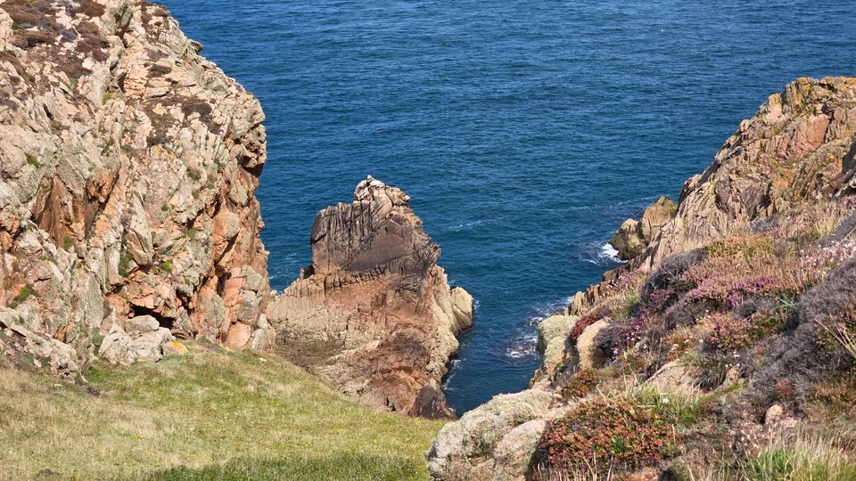 Steep rocky cliffs frame a narrow inlet and a jagged offshore rock above blue water at Grosnez.