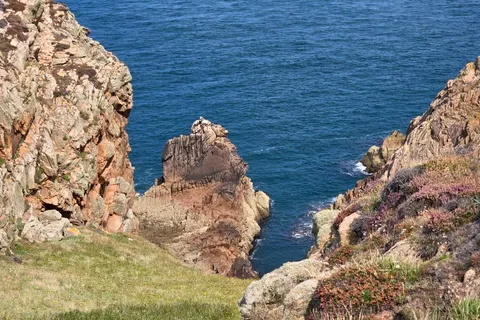 Steep rocky cliffs frame a narrow inlet and a jagged offshore rock above blue water at Grosnez.
