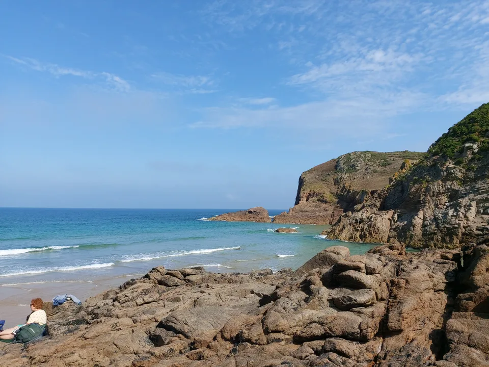 Angular coastal rocks lead towards turquoise water and a cliff at Plémont beneath a bright blue sky.