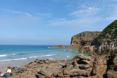 Angular coastal rocks lead towards turquoise water and a cliff at Plémont beneath a bright blue sky.