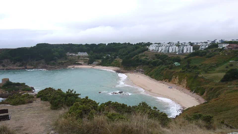 A sandy cove and rocky tidal islet lie below tree-covered slopes and white coastal buildings under a grey sky.