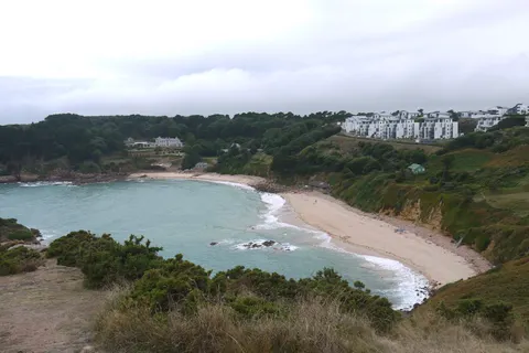 A sandy cove and rocky tidal islet lie below tree-covered slopes and white coastal buildings under a grey sky.