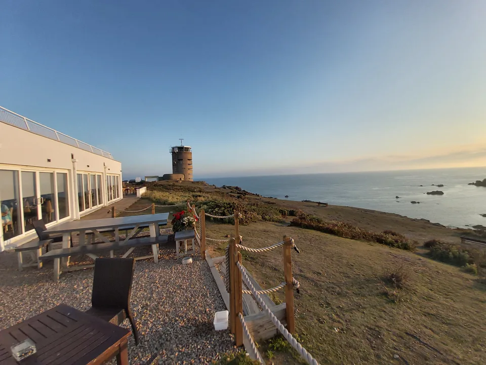 A pale building and wooden picnic tables overlook a coastal tower, grassy headland and sea at La Corbière.